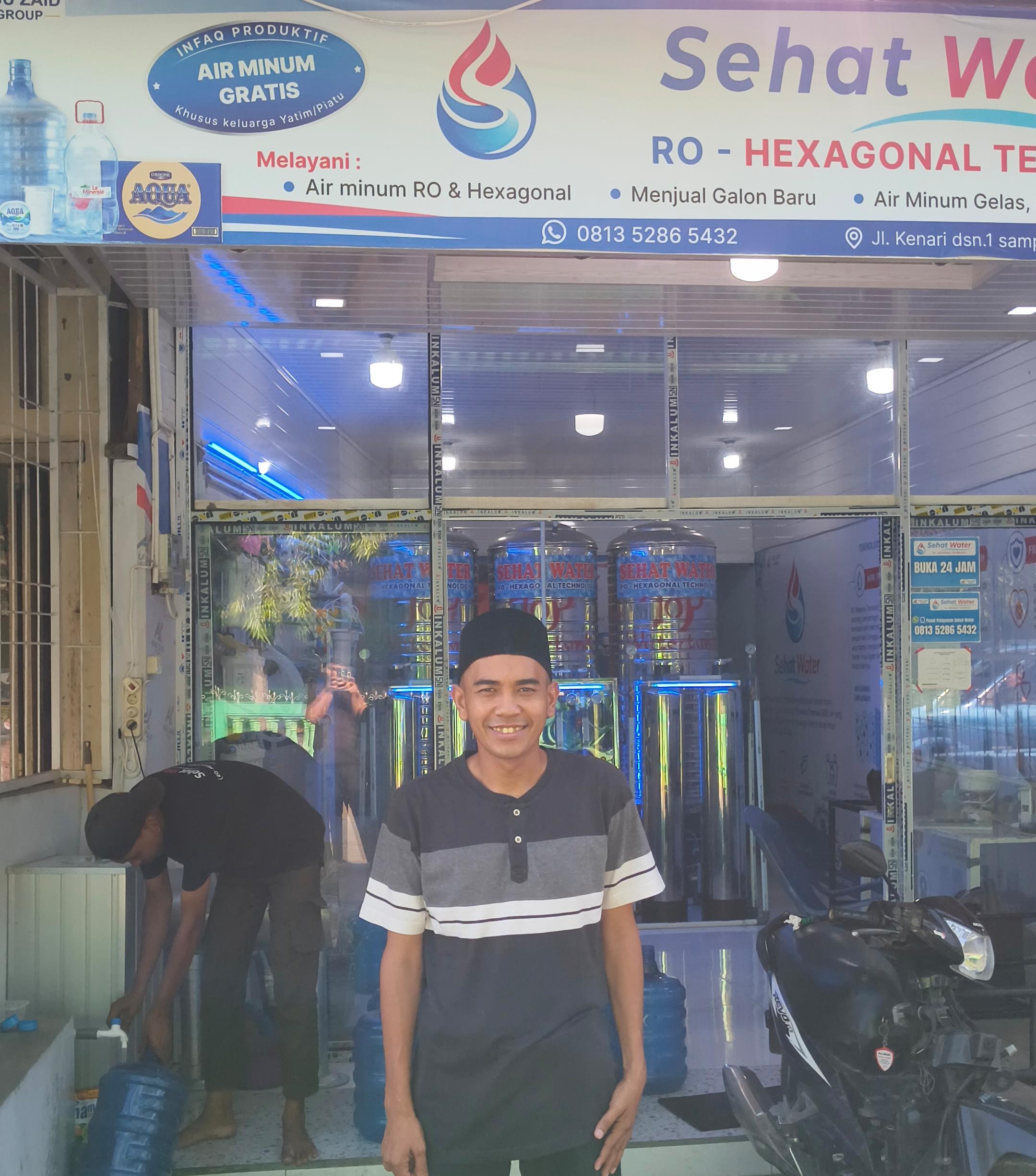 Muhammad Fajarul Aqsha pemilik depot air minum Sehat Water tersenyum di depan tokonya yang menyediakan program Infak Produktif air minum gratis bagi anak yatim piatu di Desa Kutablang, Lhokseumawe.