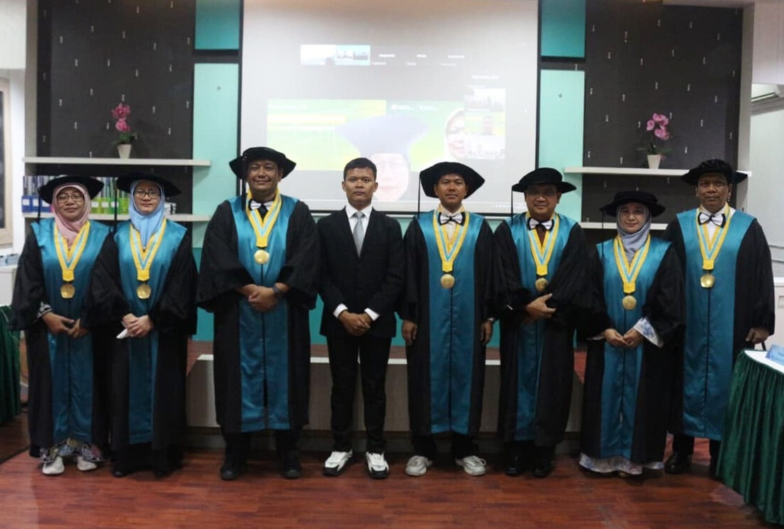 Dr. Andriy Anta Kacaribu mengenakan setelan jas hitam formal berdiri di tengah, didampingi oleh jajaran pimpinan, tim promotor, dan tim penguji Universitas Syiah Kuala yang mengenakan toga resmi pascasarjana usai Sidang Terbuka Promosi Doktor di Banda Aceh