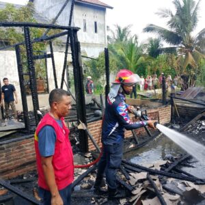 Petugas Damkar BPBD Lhokseumawe sedang menyemprotkan air menggunakan selang ke puing-puing bangunan rumah kayu yang hangus terbakar disaksikan warga.