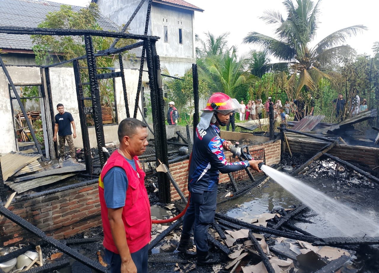 Petugas Damkar BPBD Lhokseumawe sedang menyemprotkan air menggunakan selang ke puing-puing bangunan rumah kayu yang hangus terbakar disaksikan warga.