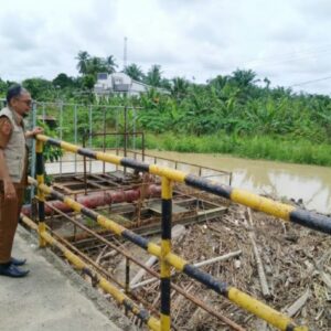 Mulyadi Kabid BPBD Aceh Utara dengan seragam dinas cokelat dan rompi krem berdiri di jembatan memantau tumpukan sampah bambu di sungai yang keruh.