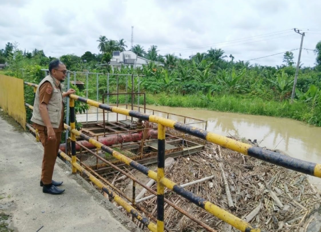 Mulyadi Kabid BPBD Aceh Utara dengan seragam dinas cokelat dan rompi krem berdiri di jembatan memantau tumpukan sampah bambu di sungai yang keruh.