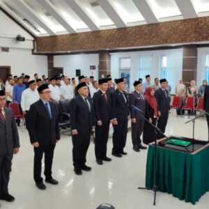 Barisan sepuluh pejabat pria dan wanita mengenakan setelan jas hitam formal dan peci berdiri tegap berjejer mendengarkan pembacaan naskah sumpah jabatan oleh bupati.