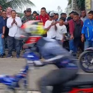 Dandim 0103 Aceh Utara Letkol Inf Faisal mengenakan kaos berkerah warna biru dongker mengangkat bendera start bermotif papan catur untuk melepas dua pembalap drag bike yang melesat cepat di lintasan balap Tanah Luas.