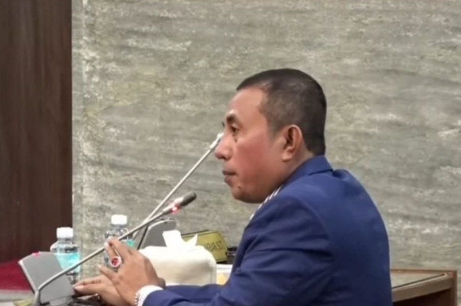 Anggota DPRA Irfannusir Rasma memakai jas biru berbicara melalui mikrofon saat melakukan interupsi dalam rapat paripurna.