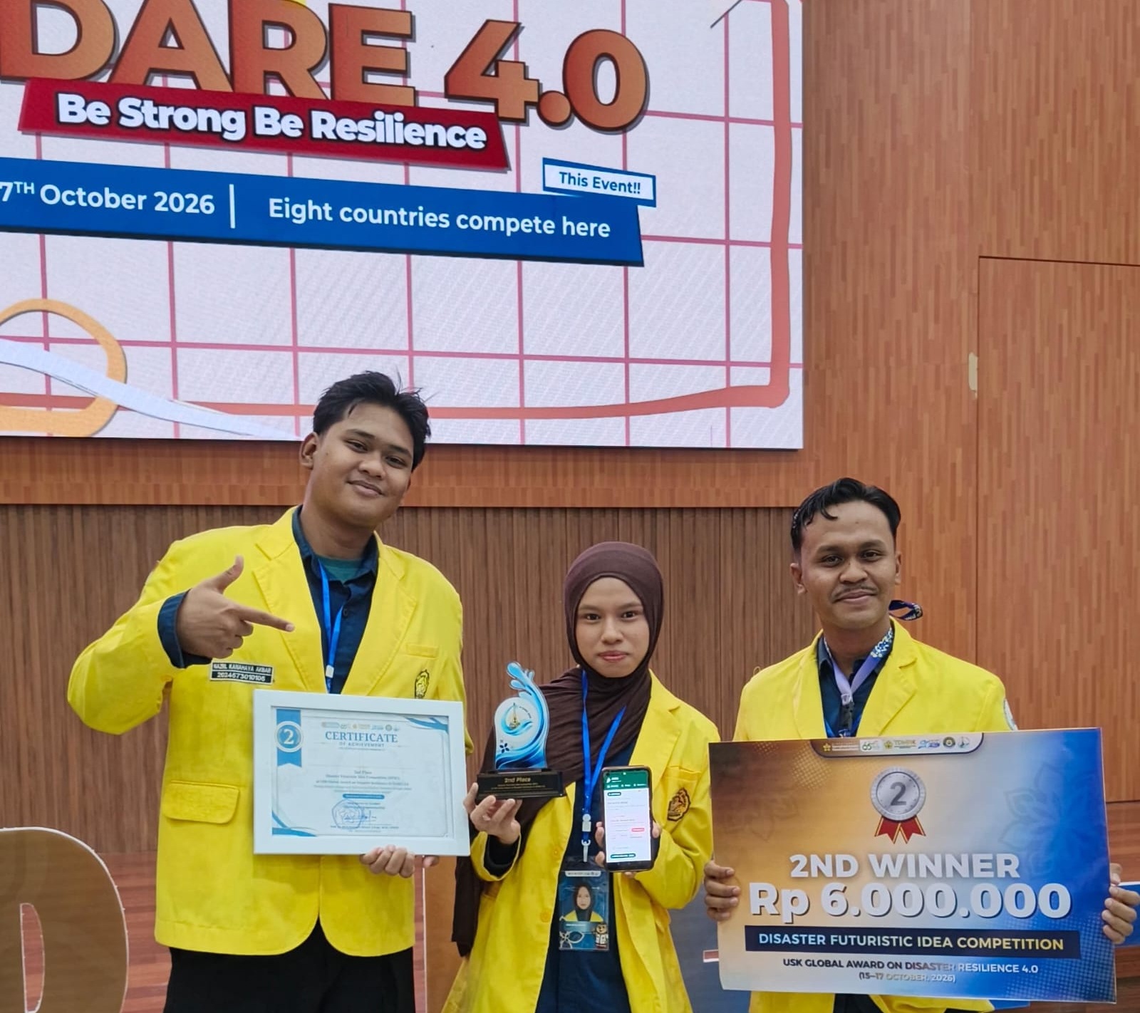 Tiga mahasiswa PNL memakai almamater kuning tersenyum bangga di atas panggung sambil memegang sertifikat, piala, handphone JAGA App, dan papan hadiah Juara 2 USK Global Award on Disaster Resilience 4.0.