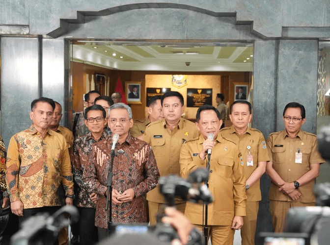 Mendagri Muhammad Tito Karnavian memegang mikrofon saat memberikan keterangan pers bersama jajaran pejabat Kemendagri dan Kemenkeu di depan pintu masuk utama berarsitektur batu alam abu-abu.