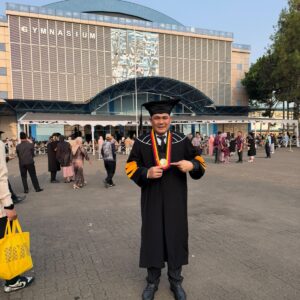 Kepala Cabang Dinas Pendidikan Wilayah Kabupaten Aceh Utara, Dr. Muhammad Johan, S.Pd., M.Pd., mengenakan toga wisuda program doktor di depan Gedung Gymnasium Universitas Pendidikan Indonesia (UPI) Bandung