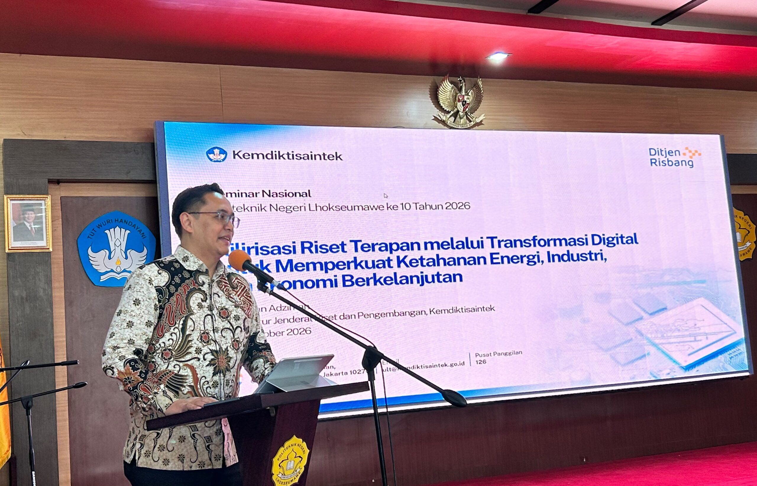 Dirjen Risbang Kemdiktisaintek Dr Fauzan Adziman memakai baju batik cokelat sedang berpidato di podium Aula PNL dengan latar belakang layar persentase digital
