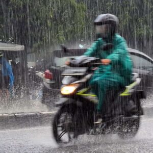 Seorang pengendara motor menggunakan jas hujan hijau menerobos hujan deras di jalan raya.