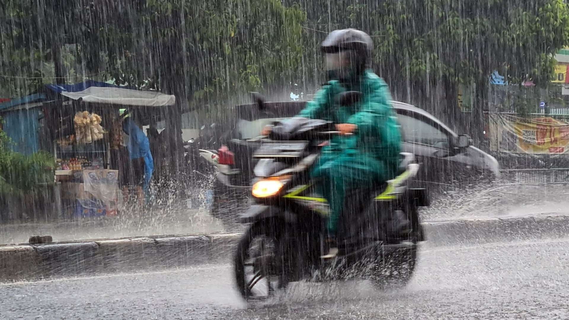 Seorang pengendara motor menggunakan jas hujan hijau menerobos hujan deras di jalan raya.