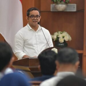 Pj. Sekda Aceh Taufik mengenakan kemeja putih dan kacamata saat menyampaikan kata sambutan di balik podium kayu dalam acara peluncuran program kemitraan demokrasi.