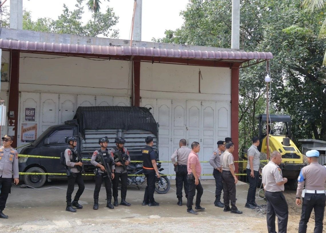 Aparat kepolisian bersenjata lengkap dan rompi antipeluru berjaga di depan dua ruko putih yang telah dipasangi garis polisi warna kuning di Gampong Beureueh Dua, Kecamatan Mutiara Timur, Pidie. Tampak sebuah mobil pikap hitam terparkir di depan ruko