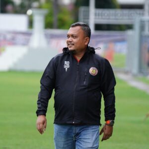 Rahmat Djailani menggunakan jaket hitam bertuliskan RD TROPS berdiri di pinggir lapangan sepak bola.