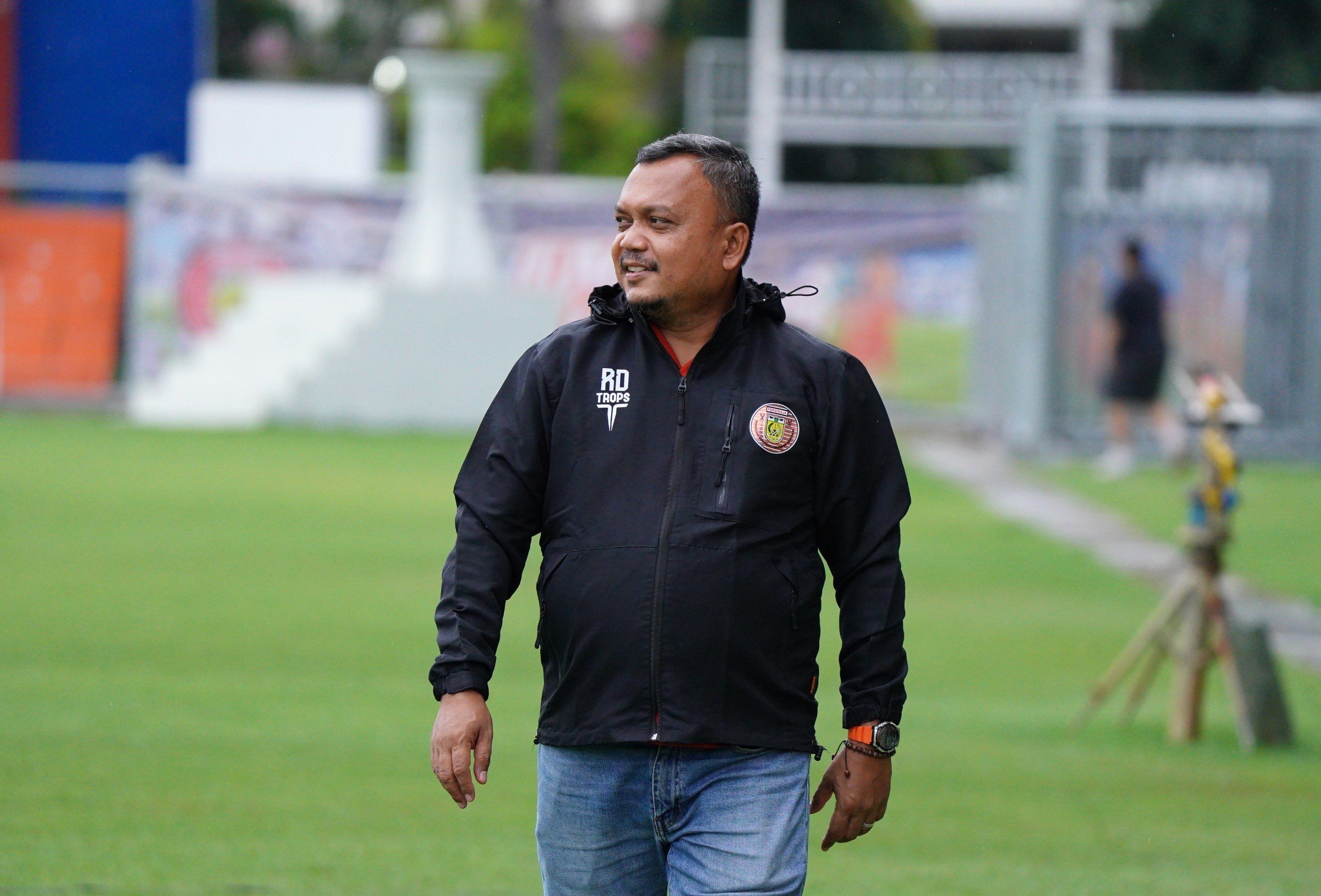 Rahmat Djailani menggunakan jaket hitam bertuliskan RD TROPS berdiri di pinggir lapangan sepak bola.